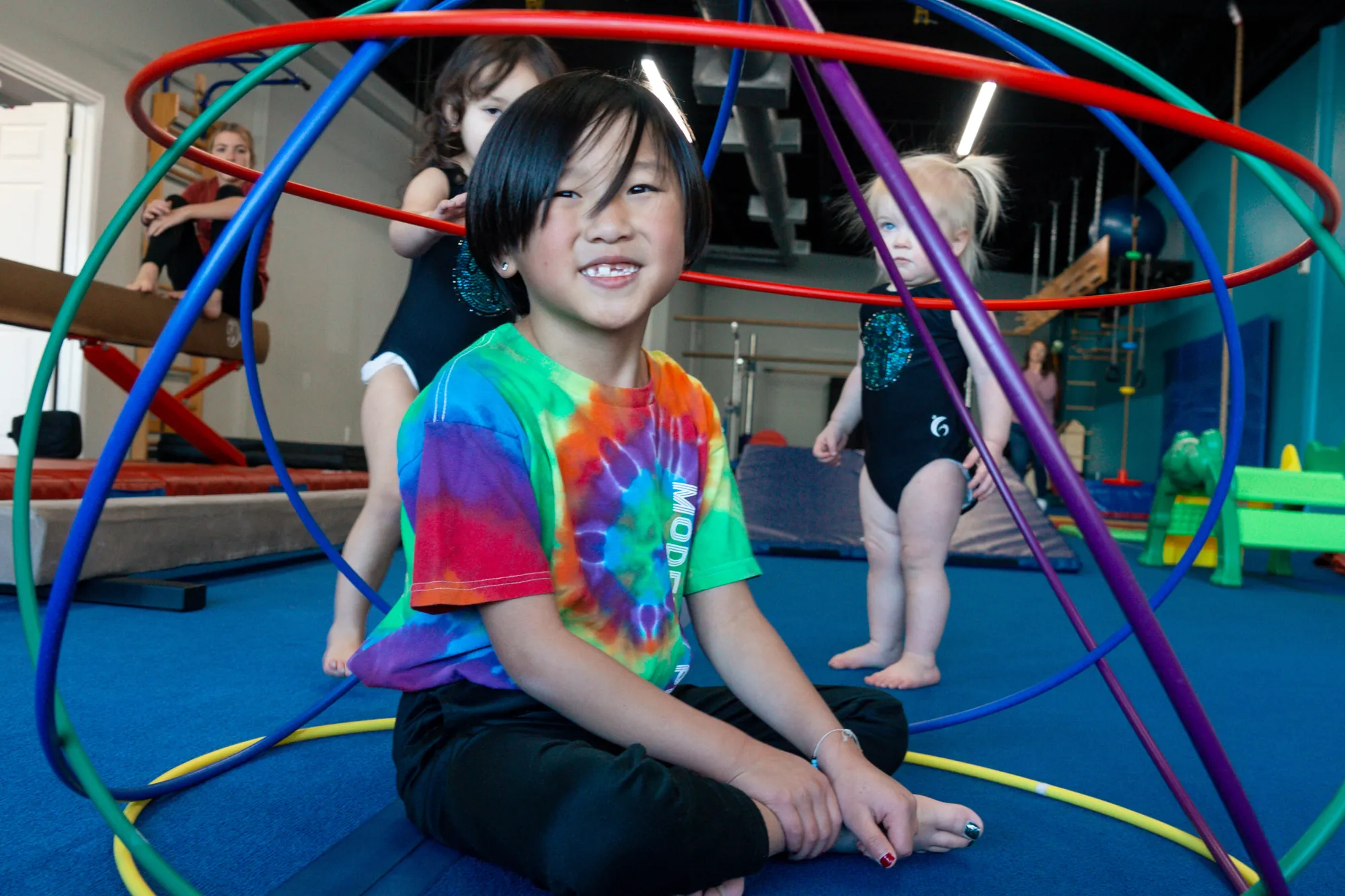A smiling kid in a tie-dye shirt sitting inside a rainbow hoop on the gym floor