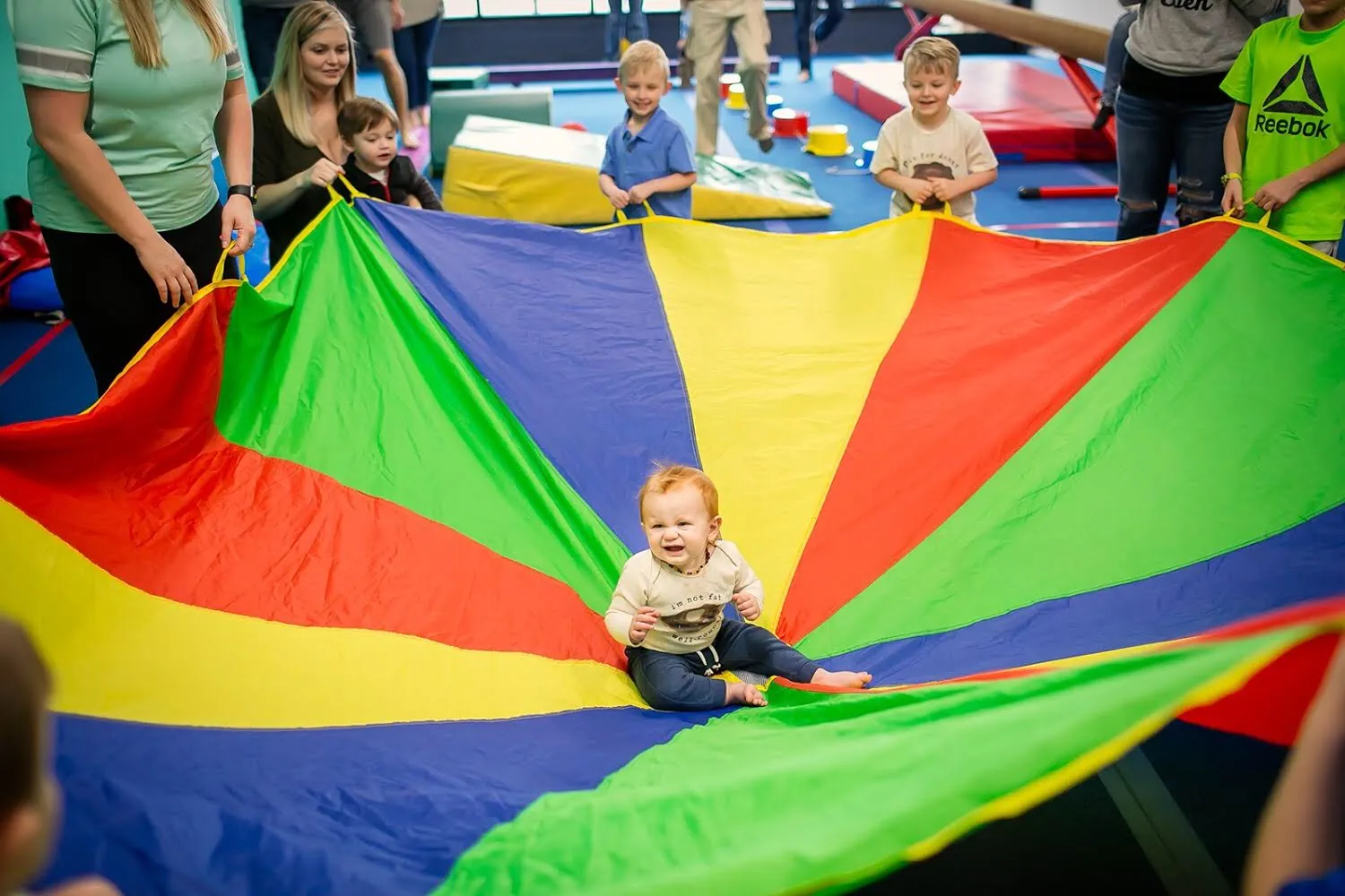 Kids and parents holding a rainbow parachute with a toddler in the middle