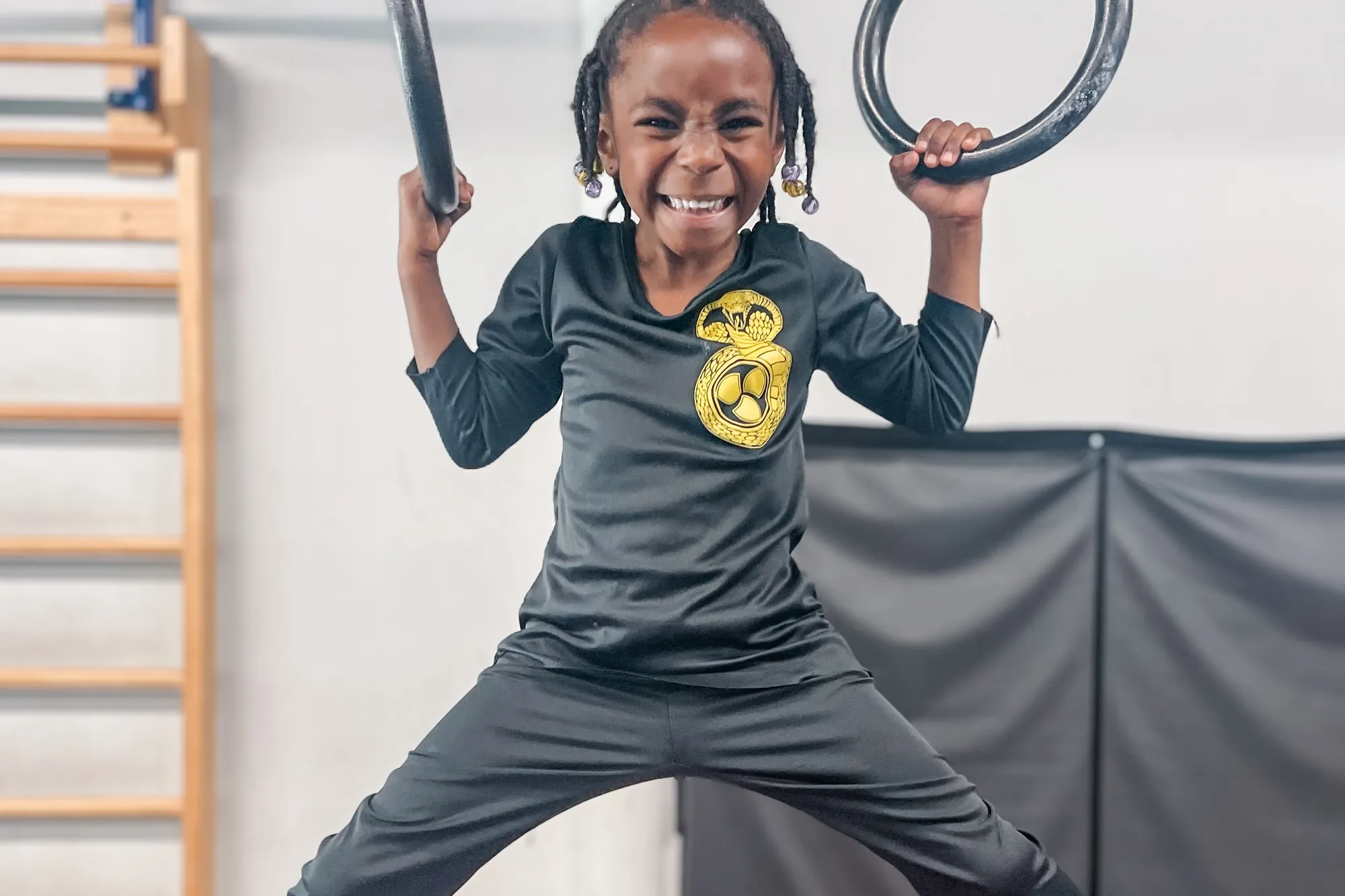 A grinning kid swinging from the gym rings with legs out wide