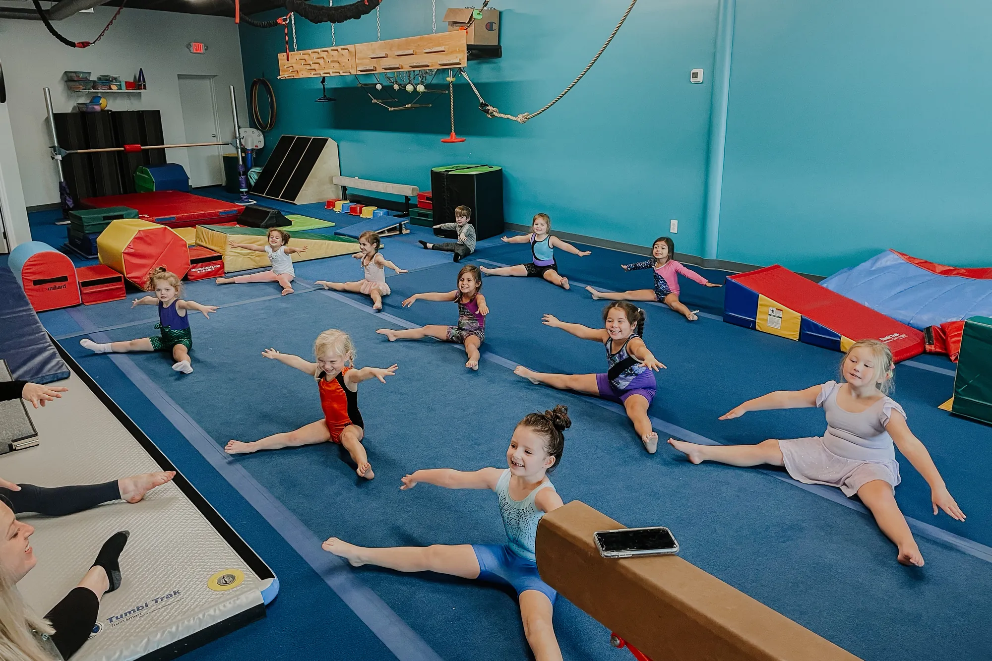 A preschool gymnastics class practicing straddle splits on the mat